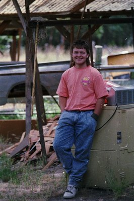 Young person in casual 1990s attire—pink graphic tee, blue jeans, and sneakers—poses outdoors under a rustic wooden shelter. ...