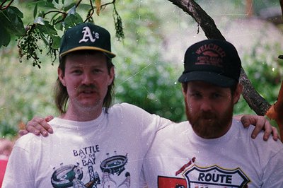 Two men pose outdoors under a tree, soaked from rain. Left wears an **A’s** cap and a **"Battle of Iwo Jima"** T-shirt; right...