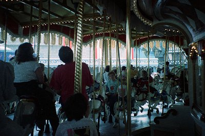 Classic mid-20th century carousel with ornate wrought-iron structure, neon-lit horses, and vintage seating. Crowded indoor am...