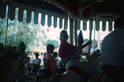 Vintage carousel with ornate, arched canopy and hanging lanterns. Operator in maroon shirt guides riders on classic wooden ho...