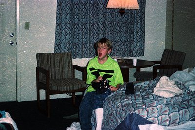 Vintage hotel room interior featuring a child in a neon-green T-shirt and jeans holding a vintage camcorder, seated on a flor...