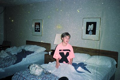 Vintage bedroom interior with twin beds, floral wallpaper, and a child in a pink shirt with a large "X" graphic. Dim lighting...