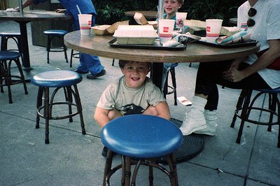 A young boy sits at a round metal table in an outdoor café, smiling at the camera. Blue vinyl stools surround him. He wears a...
