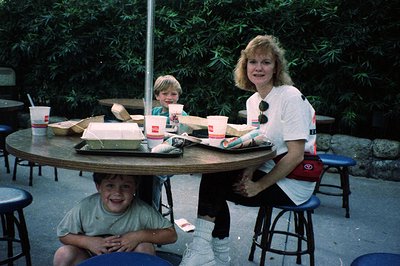 Family dining at an outdoor fast-food restaurant, likely a McDonald’s, in the 1980s–1990s. Three people—two adults and a chil...