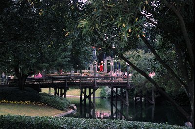 A serene park scene featuring a wooden pedestrian bridge spanning a calm pond. The bridge, supported by sturdy posts, leads t...