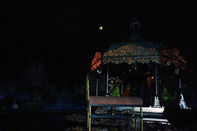 Indoor shrine adorned with ornate, draped fabric and string lights, likely for a Hindu festival. Wooden platform with offerin...