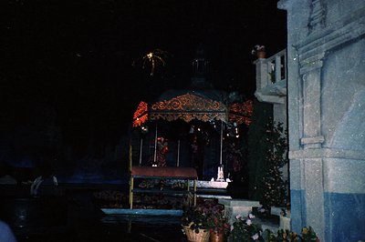 Vintage nighttime photograph of a festive courtyard pavilion adorned with red garlands and artificial flowers. A small stage ...