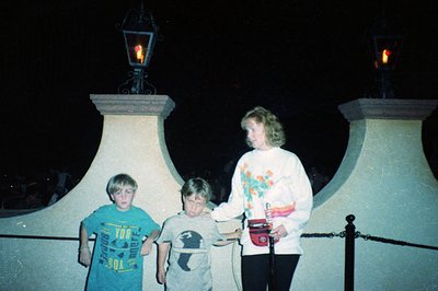 Vintage nighttime photo of three individuals posing under ornate lampposts with illuminated globes. Two children in casual 19...
