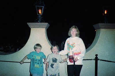 Three children and an adult pose under ornate lampposts in a dimly lit, likely amusement park setting. The adult wears a whit...