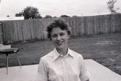 Mid-century woman in a button-down blouse poses outdoors, likely 1950s–1960s. Wooden fence and grassy yard suggest suburban r...