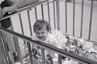 Young child in a highchair, smiling, surrounded by vintage stuffed animals. Indoor setting with metal railings and a visible ...
