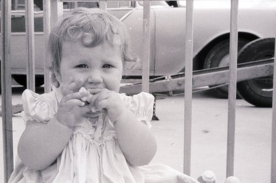 Mid-20th century black-and-white photo of a young girl in a simple white dress, covering her mouth with hands. Industrial set...