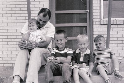 A mid-20th century family portrait on brick steps, likely 1950s–1960s. A man in a short-sleeve dress shirt holds a baby, flan...