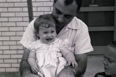 A man in a short-sleeved button-up shirt cradles a laughing toddler in a ruffled dress, mid-1960s. Brick wall and partial doo...