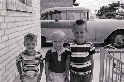 Three young boys pose outdoors in 1960s suburban America, wearing striped shirts and shorts. Classic station wagon and brick ...