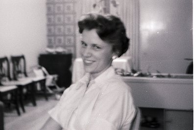 Vintage black-and-white photo of a woman in a mid-century kitchen, wearing a short-sleeved blouse and pearl earrings. Wooden ...