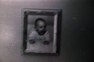 Vintage sepia-toned portrait in a distressed wooden frame, likely early 20th century. Blurred infant portrait with visible ag...
