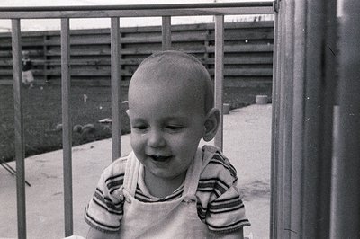 A candid black-and-white photo of a toddler laughing, leaning against a metal railing. The child wears a striped long-sleeve ...