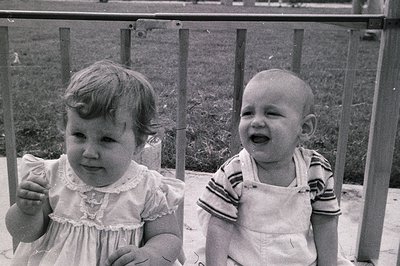 Two children in mid-20th-century attire pose on a metal playground structure. Girl in a puff-sleeve dress with lace trim, boy...