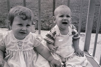 Two children sit on a concrete ledge beside metal railings, mid-20th century. Girl in a puff-sleeve dress, boy in striped ove...