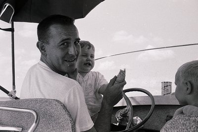 Mid-20th century family road trip in classic vintage car. Man in driver’s seat holding infant, another child visible in rear....