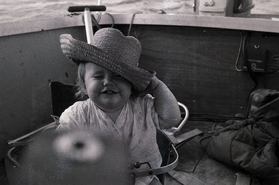 Child in vintage fishing boat, wearing wide-brimmed straw hat and light blouse, seated in a metal seat. Black-and-white, mid-...
