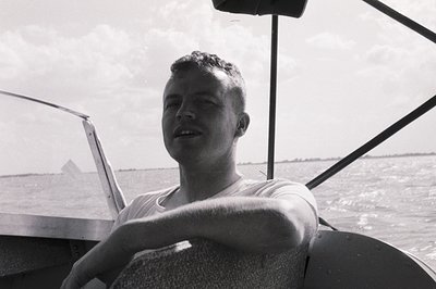 Mid-20th century man in a sleeveless naval uniform, seated at a boat’s helm with open water and distant land in background. C...