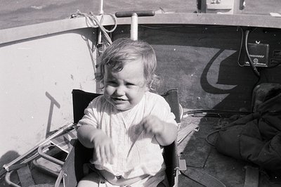 Vintage black-and-white photo of a toddler seated in a vintage boat, smiling broadly. The child wears a short-sleeved dress w...