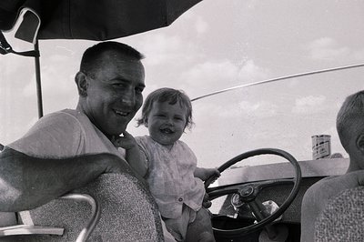 A mid-20th century black-and-white photo captures a man and child in an open-air vintage car, likely a 1950s–1960s model. The...