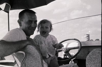 Black-and-white candid of a man and young child laughing in an open-air vintage vehicle, likely a 1950s–1960s American sedan....