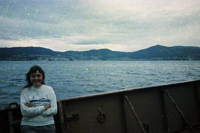 Vintage polaroid-style shot of a young woman standing on a weathered boat deck, holding a camera. She wears a light sweater w...