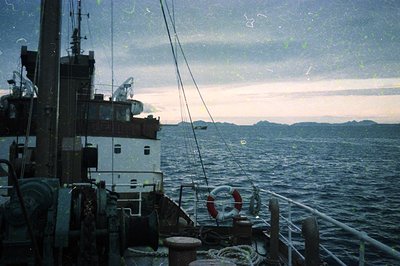 Vintage fishing vessel deck with worn metal railings, lifebuoy, and coiled ropes against a misty coastal horizon. Distressed ...