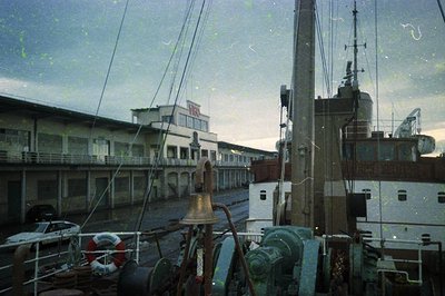Vintage maritime scene featuring a dockside industrial port. Prominent rusted ship’s bell and lifebuoys in foreground. Mid-20...