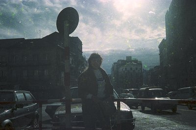 Urban street scene viewed through rain-streaked glass, showing a lone figure in a dark coat and hat standing near a pole. Mid...