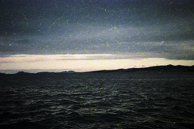 Milky Way galaxy arcs over a dark ocean horizon, silhouetted by faint moonlight. Storm clouds and distant landforms create co...
