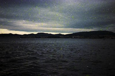 Silhouetted mountain range under dramatic night sky with faint aurora-like lights over dark waters. Moody, atmospheric seasca...