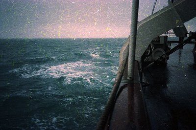 Vintage film grain captures rough seas from a ship’s rail, with choppy waves and misty horizon. Rustic metal railing and blur...