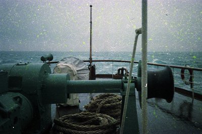 Mid-20th century naval bridge deck in heavy seas, featuring a green-painted metal railing, coiled ropes, and a covered radar/...