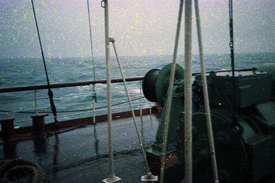 Vintage ship deck in rough seas, featuring rusted metal railings, a green winch, and taut ropes. Raindrops blur the scene, em...
