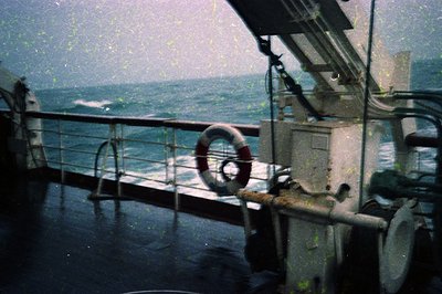Vintage ship deck view through rain-streaked glass, showing a lifebuoy mounted on metal railing. Industrial maritime equipmen...