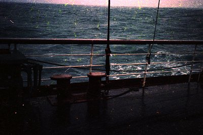 Midship deck view through railings showing turbulent sea. Rusty mooring bollards and taut ropes suggest maritime setting. Low...
