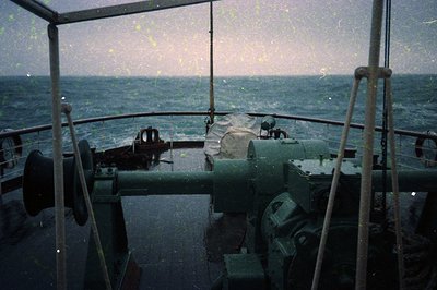 Vintage ship bridge interior with rain-streaked windows, showing open sea beyond. Rusty metal railings, control wheel, and na...