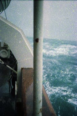 Vintage view through a ship’s porthole showing turbulent seas and stormy horizon. Rusty metal framing suggests mid-20th centu...