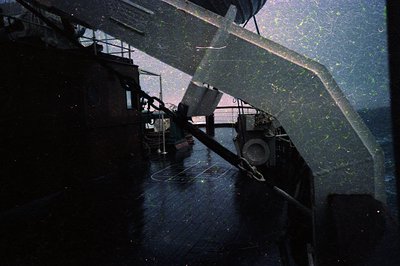 Industrial ship deck with angled perspective, showing rusted metal railings, cargo winches, and a lone worker in dark clothin...