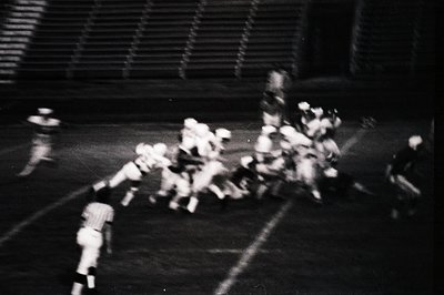Blurred black-and-white shot of a dynamic football (soccer) match in progress, likely mid-1970s. Players in motion on a grass...