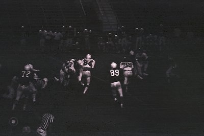 Black-and-white shot of an indoor football game, likely from the 1960s–70s. Players in numbered jerseys (, ) engage in a scru...