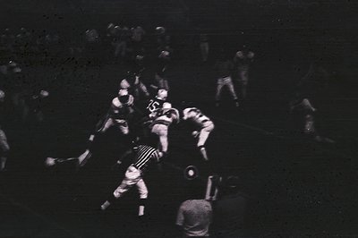 Vintage black-and-white photo capturing a chaotic football huddle in low-light stadium. Players in striped jerseys and helmet...