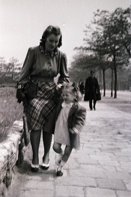 A woman in a 1950s-style pleated skirt and cardigan leads a young girl by the hand along a tree-lined path. The woman’s plaid...