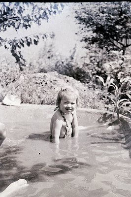 Mid-20th century black-and-white photo of a toddler in a shallow pool, wearing a woven belt. Lush foliage and stone walls fra...