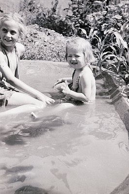 Two young girls sit in a shallow outdoor pool, surrounded by lush greenery. The older girl, wearing a one-piece swimsuit, smi...